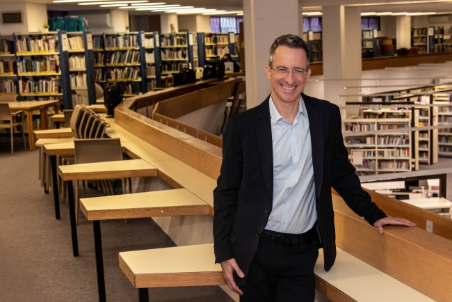 Centenary University psychology professor Tal Ben-Shahar, a light-skinned man with short hair wearing glasses, stands in front of shelves of books.